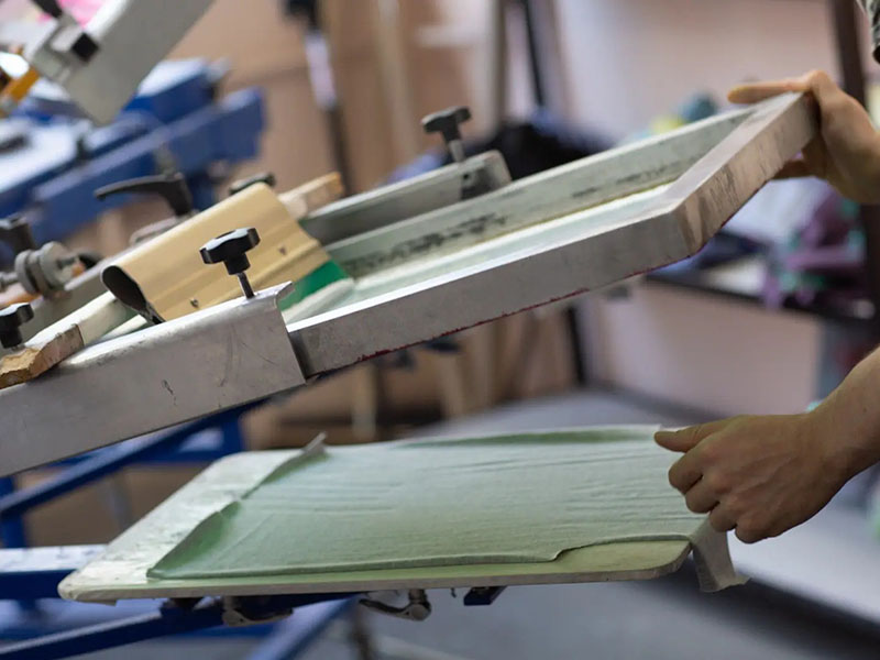 Worker lifting screen printing frame over green fabric in workshop