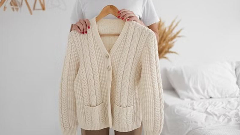 Person holding a cream-colored knitted cardigan on a hanger. The cardigan features a cable knit pattern