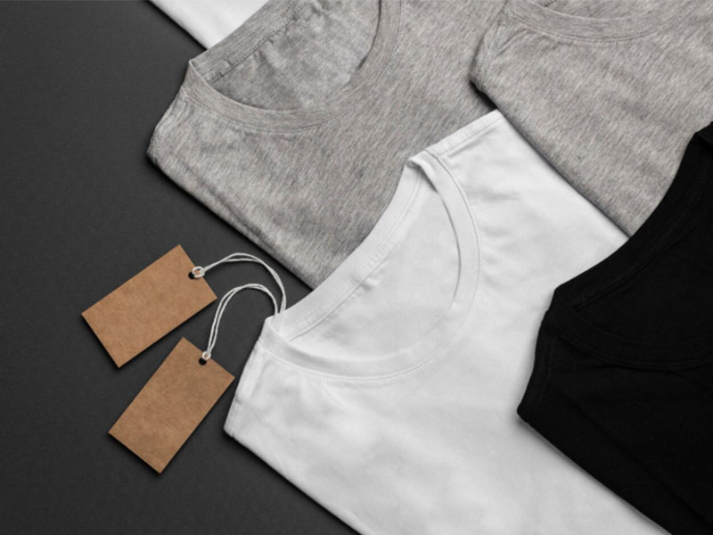 Four folded T-shirts in grey, white, and black, each with a blank brown tag attached