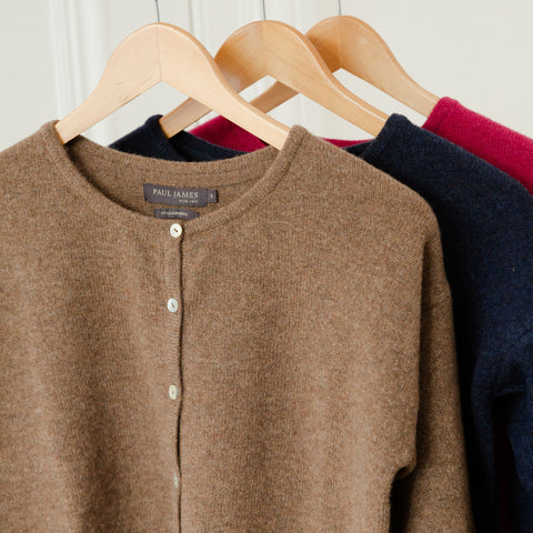 Three cardigans hanging on wooden hangers. The front cardigan is brown with a round neckline and white buttons, labeled "Paul James." Behind it are a navy and a magenta cardigan.