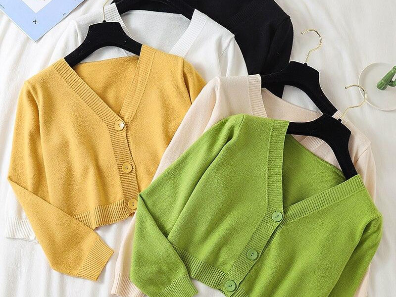 Five V-neck cardigans in different colors (yellow, green, cream, white, black) neatly arranged on black hangers on a flat surface.