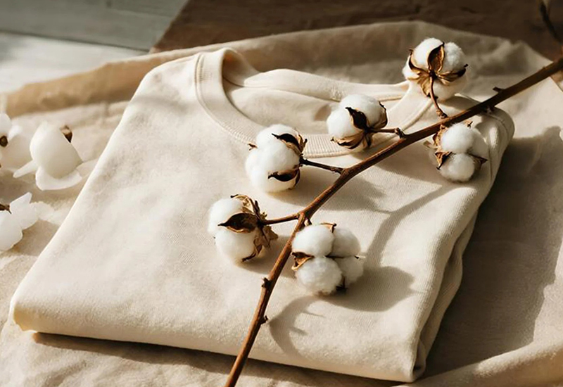 Folded cream sweatshirt styled with cotton branches on soft neutral fabric background