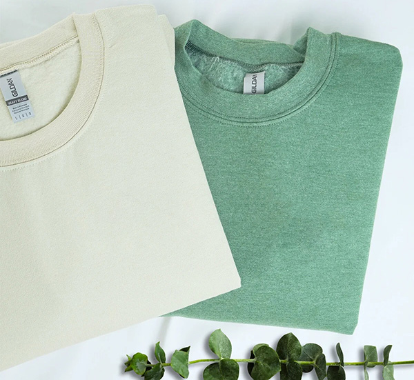 Two folded crewneck sweatshirts in cream and heather green laid flat with eucalyptus leaves.