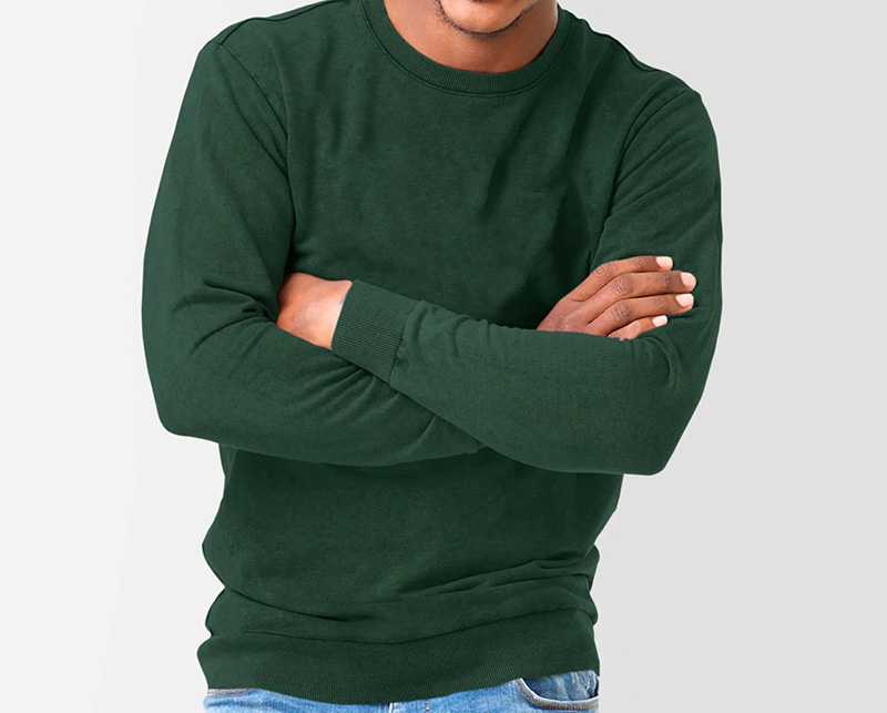 Man wearing dark green crewneck sweatshirt with folded arms on plain background.
