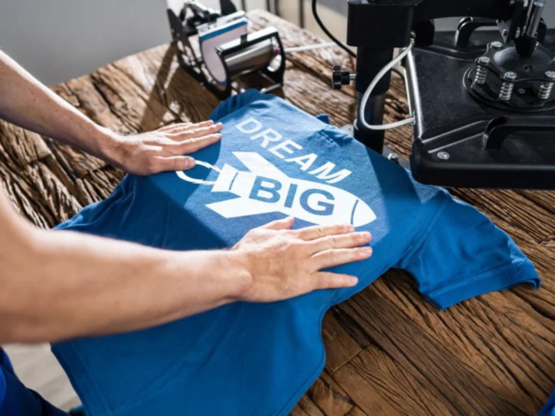 Hands positioning blue printed T-shirt under heat press machine on wooden table
