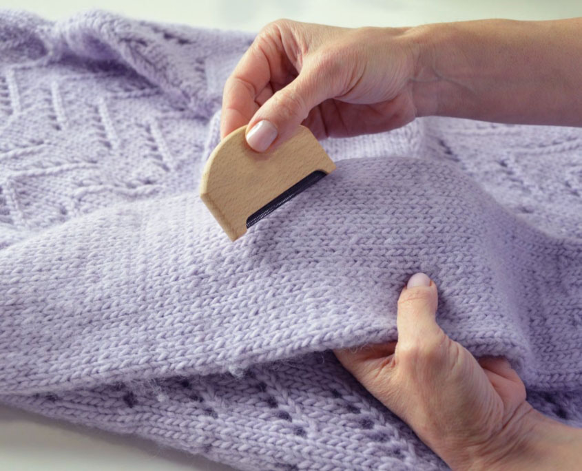 Hands using a small wooden sweater comb to remove pills from a lavender knit sweater.
