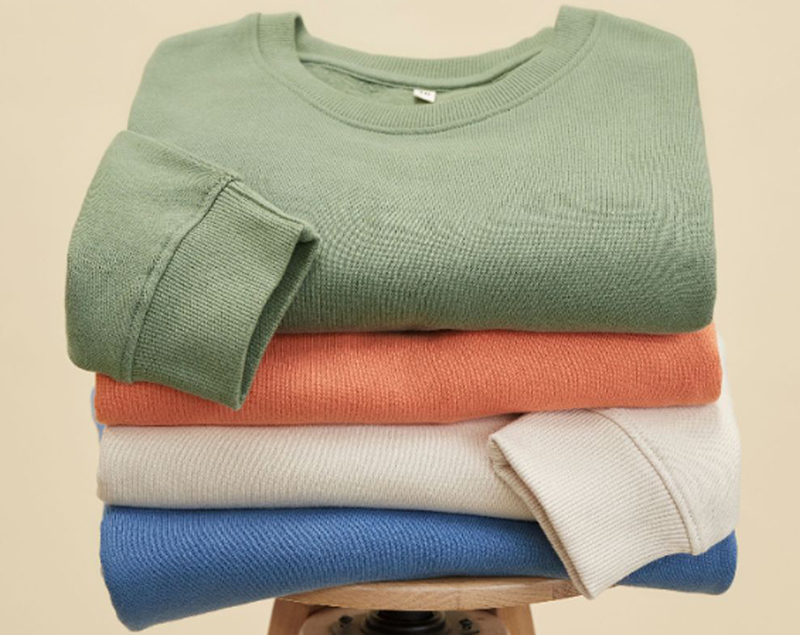 Stack of folded knit sweatshirts in sage green, coral, cream, and blue tones