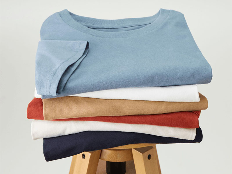 A stack of neatly folded T-shirts in various colors placed on a wooden stool against a plain background.