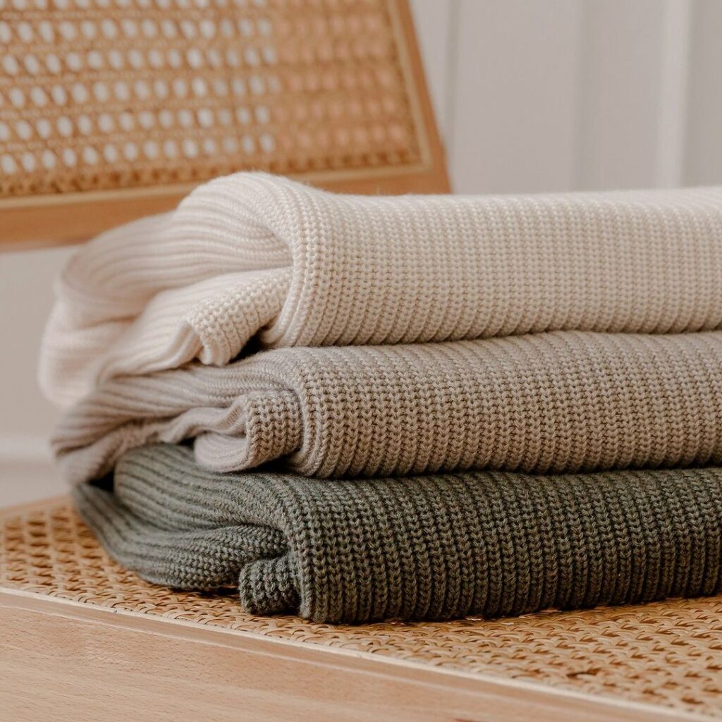 Three neatly folded knit sweaters stacked on a woven wooden chair. The top sweater is off-white, the middle is light beige, and the bottom is olive green.