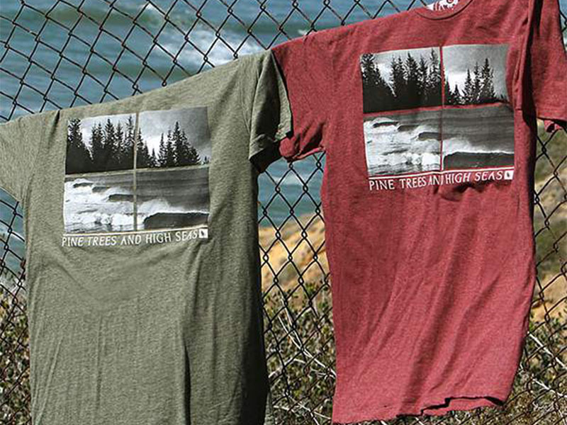 Two graphic T-shirts, green and red, hanging on chain-link fence by the ocean