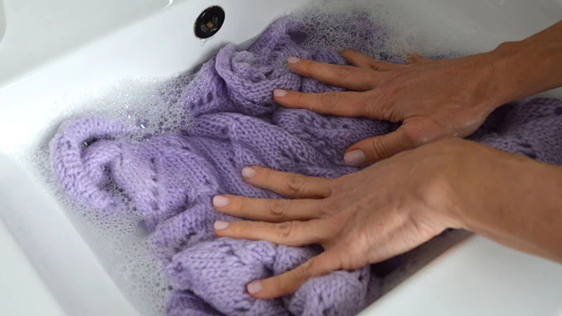 Hands gently hand-washing a lavender knit cardigan in soapy wate