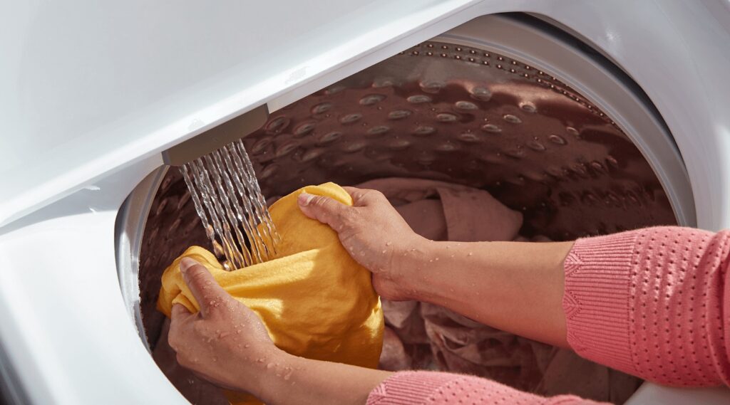 Hands washing a yellow garment inside a front-loading washing machine.