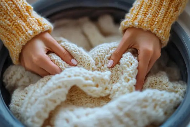 Hands holding a chunky cream knit sweater in a wash basin for delicate hand washing.