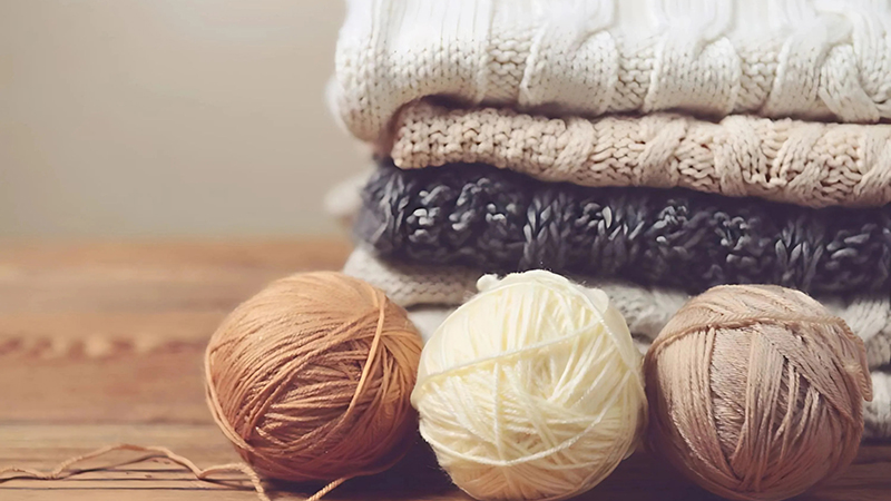 Stacked knit sweaters with yarn balls arranged on wooden table in soft neutral tones