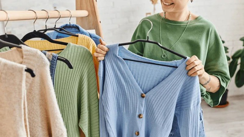 Person holding a light blue ribbed cardigan while browsing colorful knitwear on a clothing rack