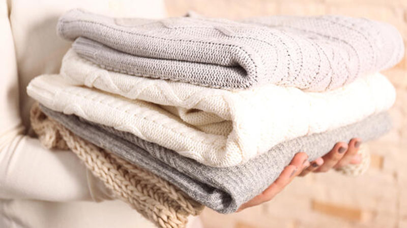 A person holding a stack of folded knitted sweaters in neutral colors, including beige, white, gray, and light brown.
