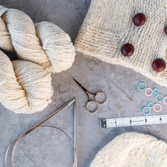 Knitting supplies with cream yarn, needles, scissors, measuring tape, and textured cardigan sample