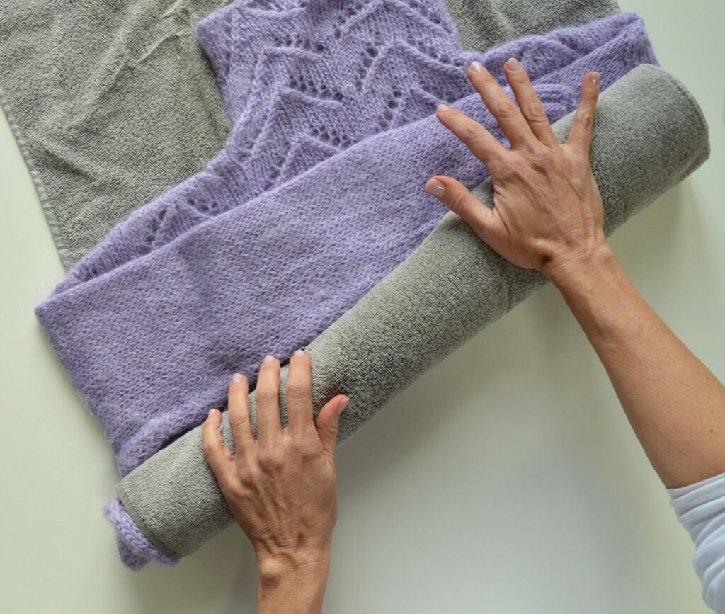 A person’s hands rolling up a knitted lavender fabric over a gray towel, showing texture and thickness.