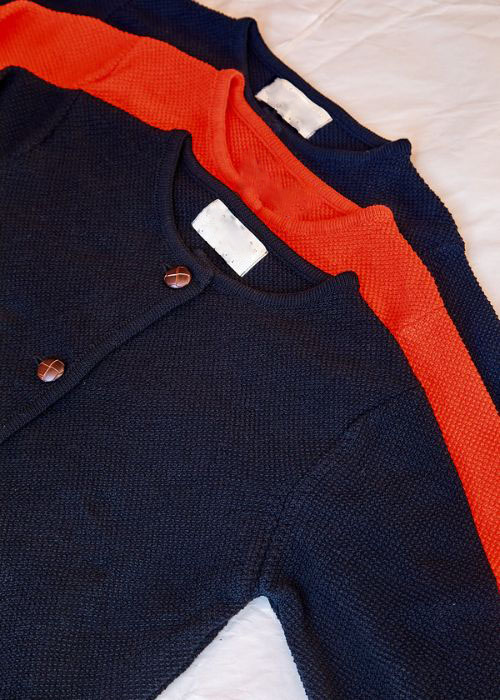Three folded cardigans, two navy blue and one bright orange, neatly stacked on a white surface, showing the textured knit fabric and front button details.
