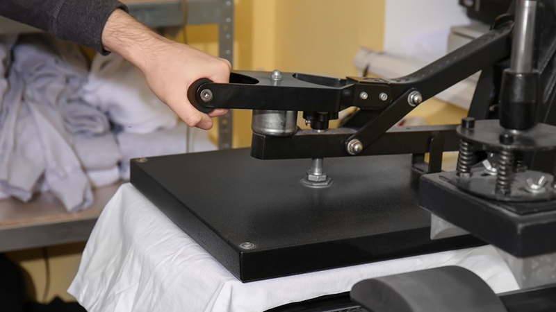 Hand operating heat press machine on white T-shirt in workshop