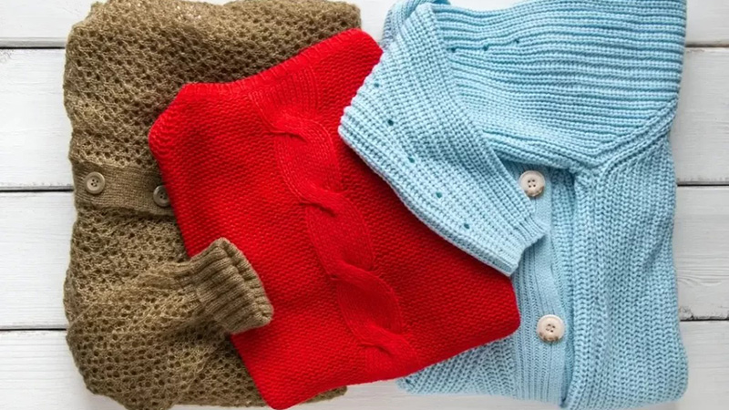 Folded knit sweaters in brown, red, and light blue arranged on white wooden background