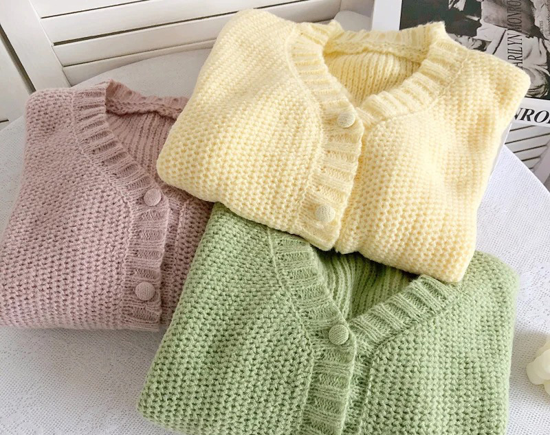 Three pastel knit cardigans folded neatly in blush pink, pale yellow, and soft green.