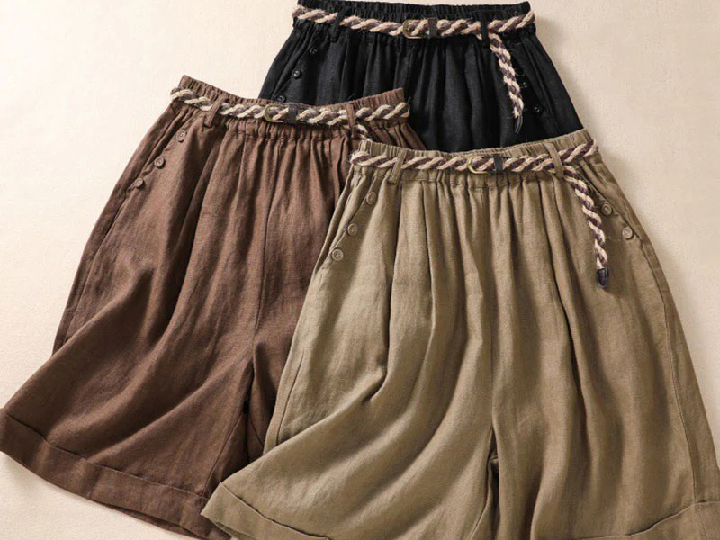 Three pairs of loose, wide-leg shorts arranged on a flat surface. The shorts are in black, brown, and olive colors, each featuring a braided rope belt and button details near the waistband.