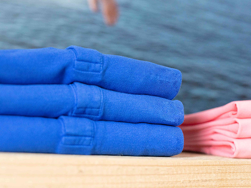 Close-up of neatly folded blue and pink polo shirts stacked on a wooden surface with a blurred water background. The shirts are made of a textured knit fabric.