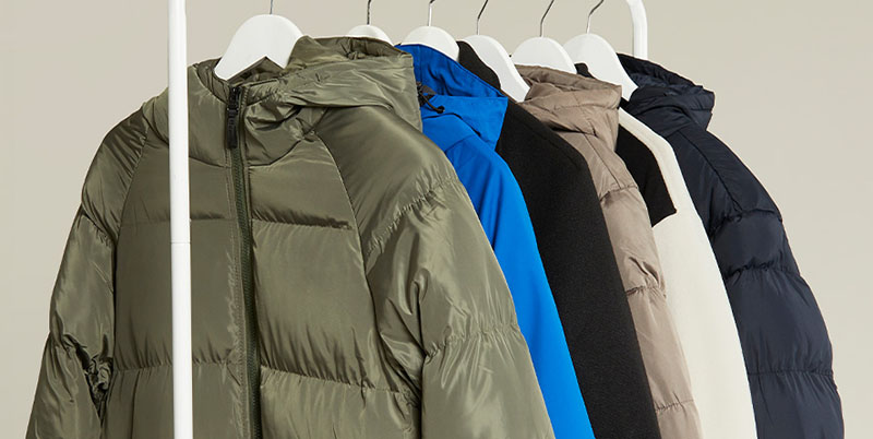 A row of hooded jackets in green, blue, beige, white, and black, hanging neatly on white hangers against a neutral background.