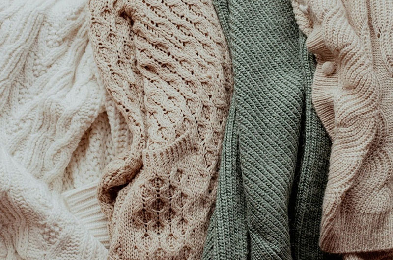 Close-up of several folded knit sweaters in cream, beige, green, and light brown, showing different textured patterns.