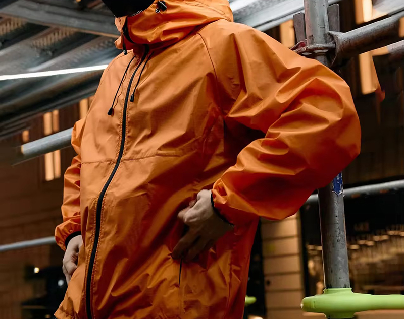 Person wearing a bright orange waterproof jacket with a hood and black zipper, standing outdoors near scaffolding, hands in the side pockets, showing a slightly relaxed fit.