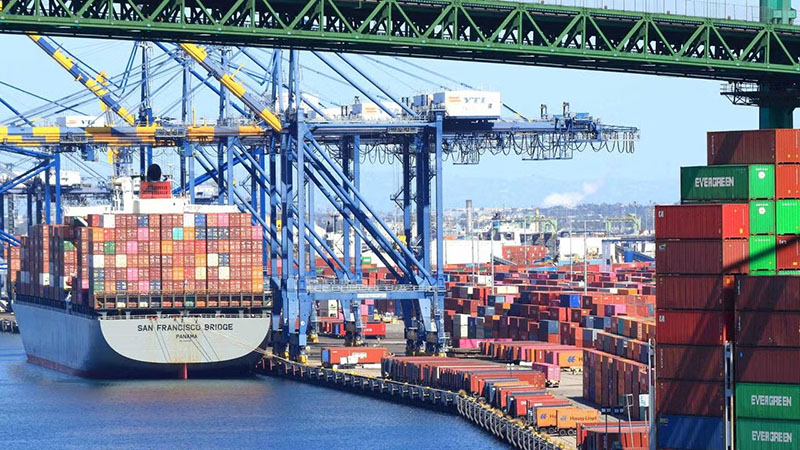 Large cargo ship named "San Francisco Bridge" docked at a busy port, surrounded by colorful shipping containers and blue cranes loading and unloading cargo.