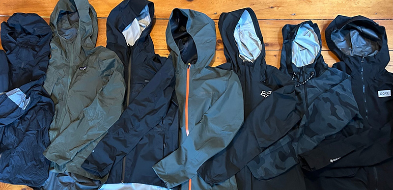 A row of seven outdoor jackets in shades of black, dark green, and camo, all with hoods, laid out side by side on a wooden floor.