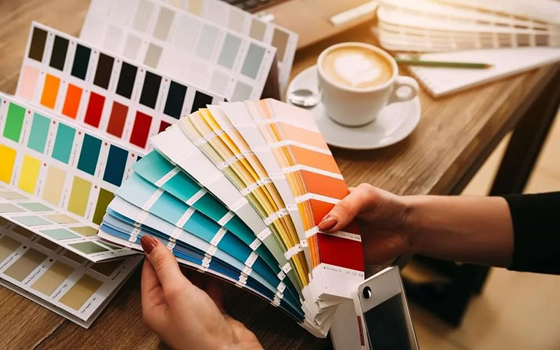 Person holding multiple color swatches and fan decks on a table with coffee cup.