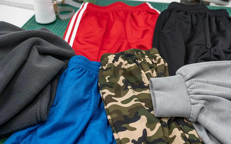 A variety of athletic pants including red, black, blue, camouflage, and gray, laid out on a green cutting mat with fabric and sewing supplies nearby.