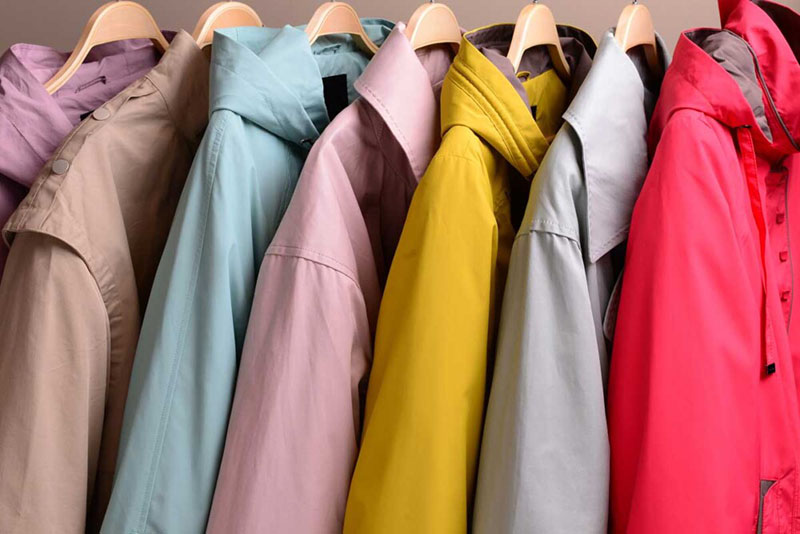 Seven rain jackets in pastel and bright colors including lavender, beige, light blue, pink, yellow, gray, and red hanging on wooden hangers in a row.