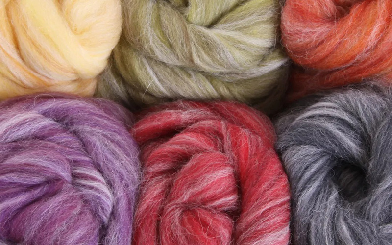 Six tightly coiled bundles of wool roving in different colors including yellow, green, orange, purple, red, and gray, arranged closely together. The wool has a soft, fluffy texture with visible fibers.