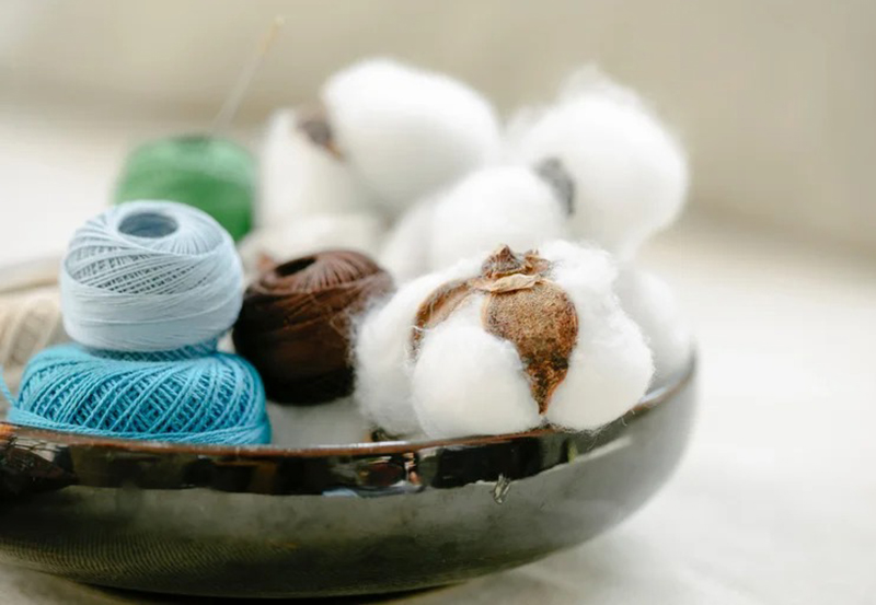 A shallow dark bowl holding several cotton bolls alongside spools of thread in various colors including blue, green, brown, and light blue.
