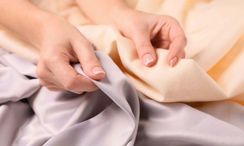 Two hands gently holding and feeling soft fabric in pastel shades of lavender and light beige, highlighting texture and smoothness.