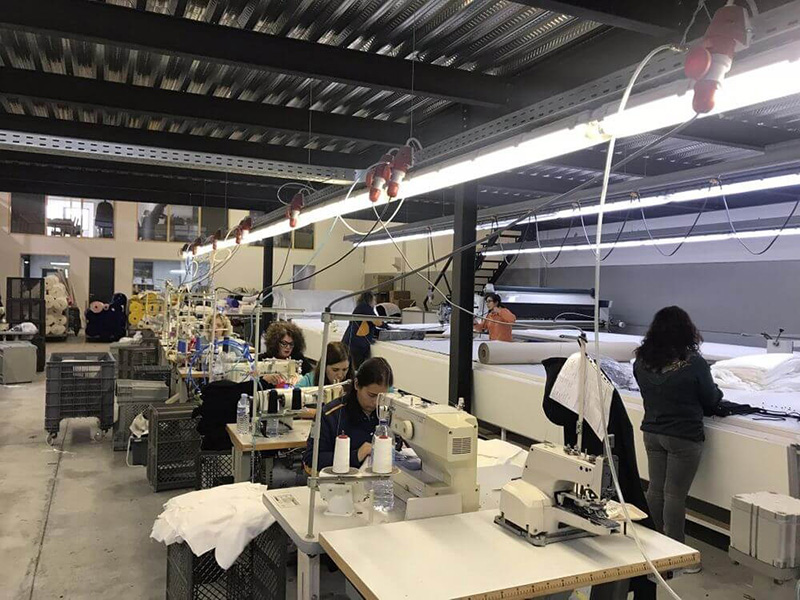 Workers operating sewing machines in a textile factory, stitching white fabric on multiple workstations under bright overhead lighting.