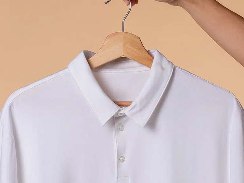 A close-up of a plain white polo shirt on a wooden hanger, held by a hand against a beige background.