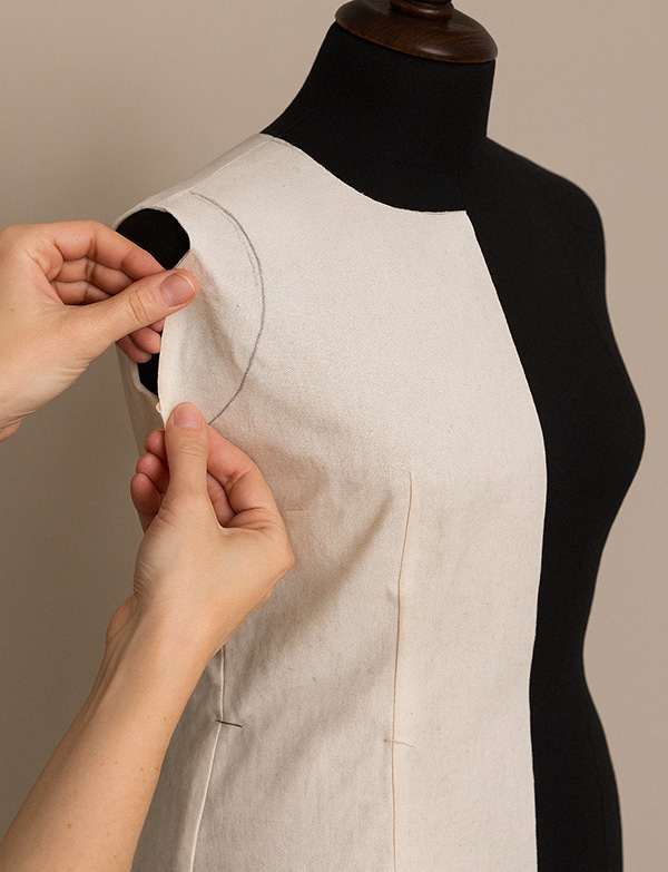 Close-up of a dress form with a muslin fabric pinned and draped on it, while a person holds and shapes the fabric around the armhole.