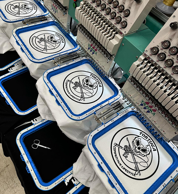 Multiple embroidery machines stitching skull logo designs onto fabric in production line.