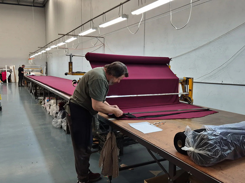 A worker in a textile factory is carefully measuring and cutting deep purple fabric on a long cutting table.