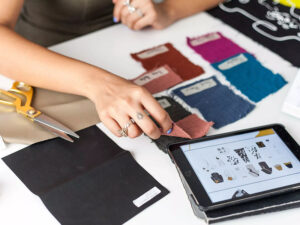 Designer reviewing fabric swatches and digital clothing tech pack on tablet workspace desk
