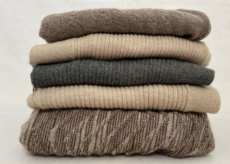 A neatly stacked pile of five folded knit sweaters in neutral colors, ranging from brown, beige, and gray tones, on a light surface.