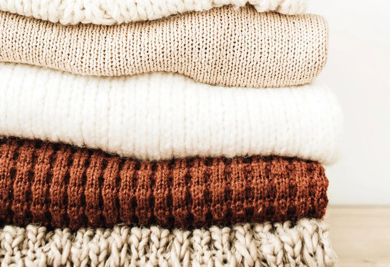 Close-up view of a neat stack of four folded knitted sweaters in shades of cream, white, brown, and beige, showing the texture of the knit patterns.