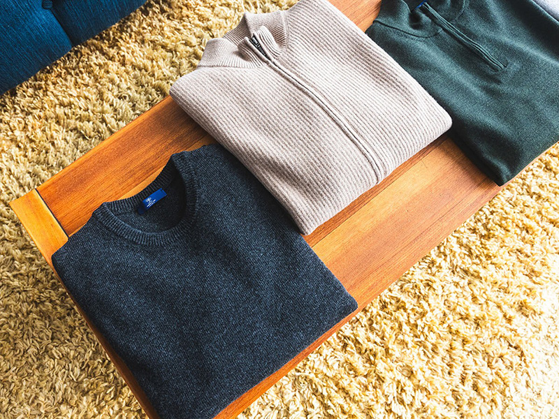 hree neatly folded sweaters, one dark gray, one beige zip-up, and one forest green, are displayed on a wooden bench over a shaggy beige carpet. The sweaters are arranged side by side, showing their textures and colors.