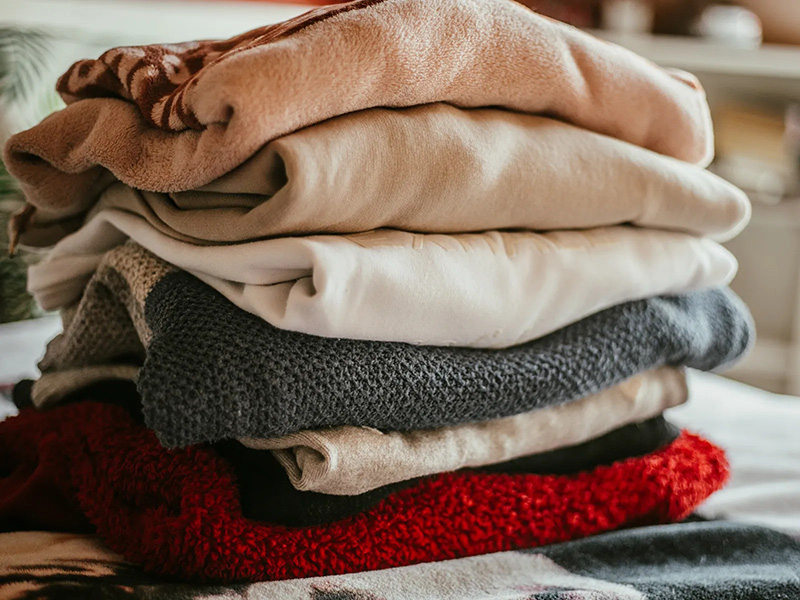 Stack of folded sweaters and soft tops in neutral tones with a bright red knit layer.