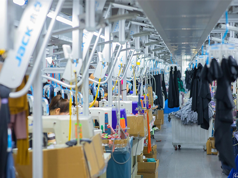 Long row of industrial sewing machines in a garment factory, workers seated at machines, clothes hanging overhead and on racks, bright lighting, with threads and materials visible on tables.